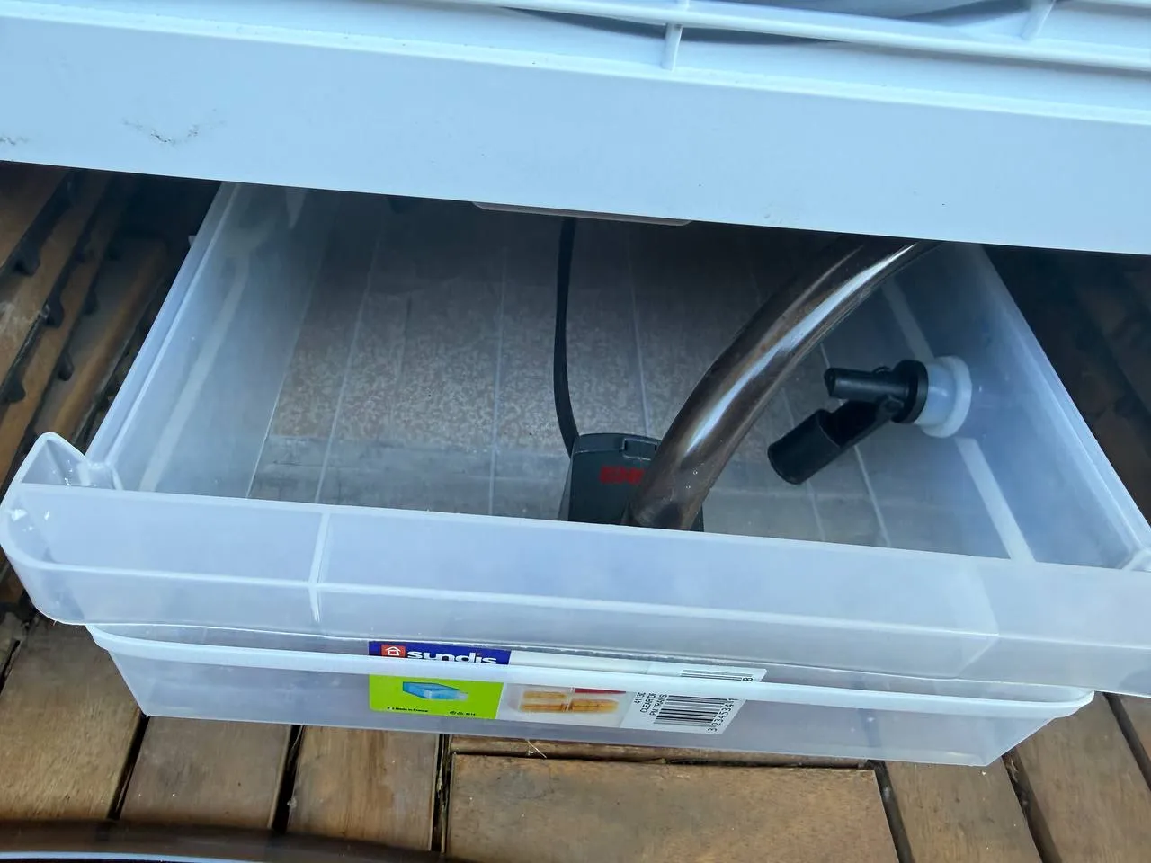 Gros plan sur l’intérieur du bac : pompe EHEIM posée au fond, câble noir d’alimentation et tuyau souple transparent raccordé à un coude noir et blanc.