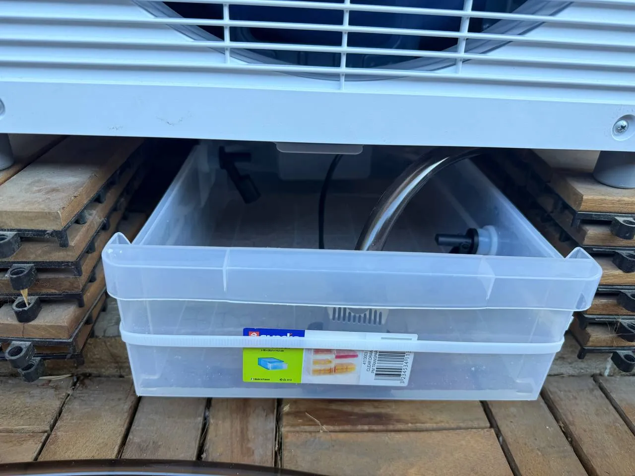 Bac plastique transparent placé sous la grille de ventilation du climatiseur, laissant voir la pompe et le tuyau à l’intérieur.