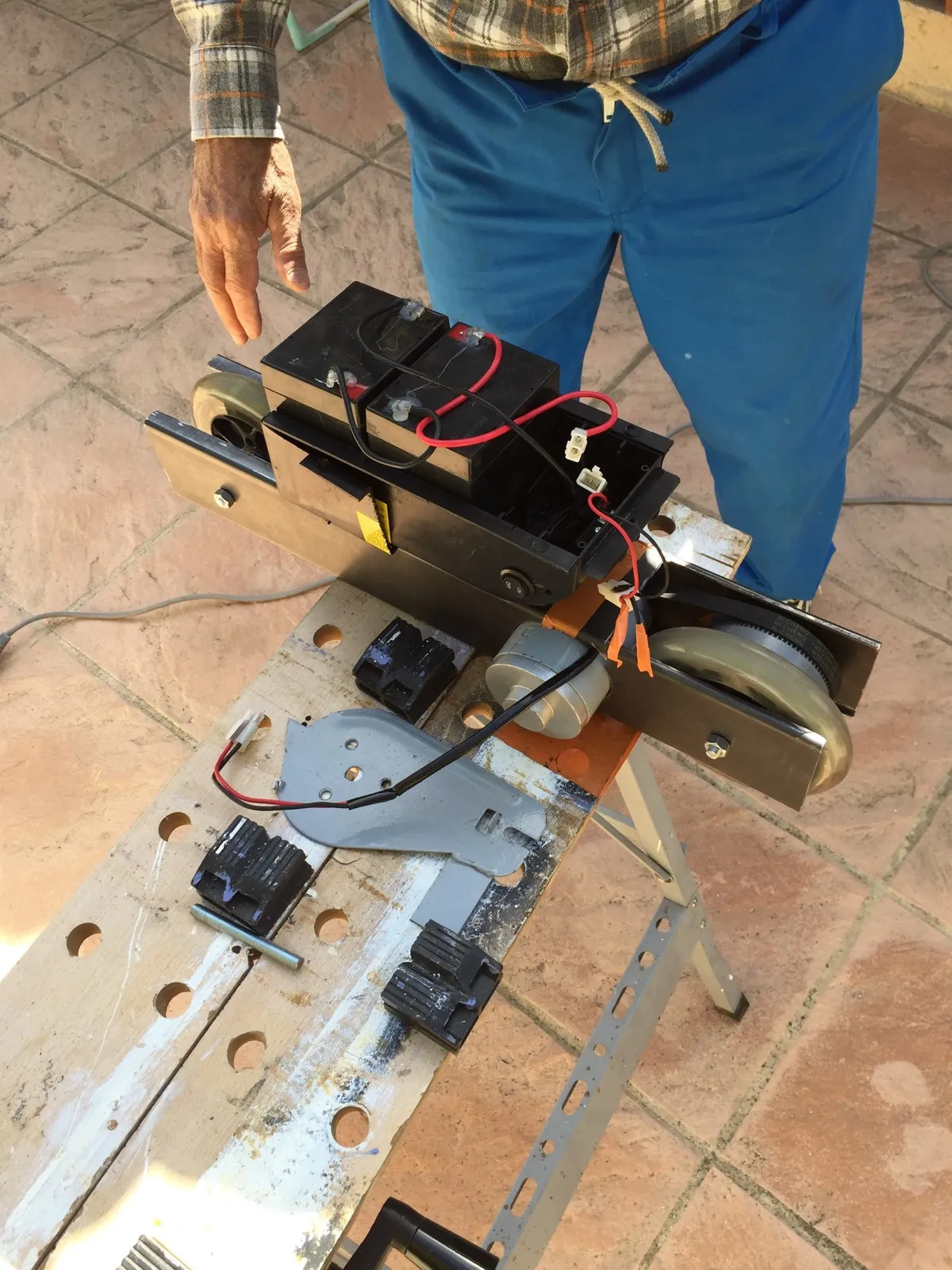 Ensemble mécanique posé sur un tréteau : cadre en aluminium, moteur cylindrique relié à une poulie crantée par une courroie, deux batteries plomb noires câblées en rouge et noir.