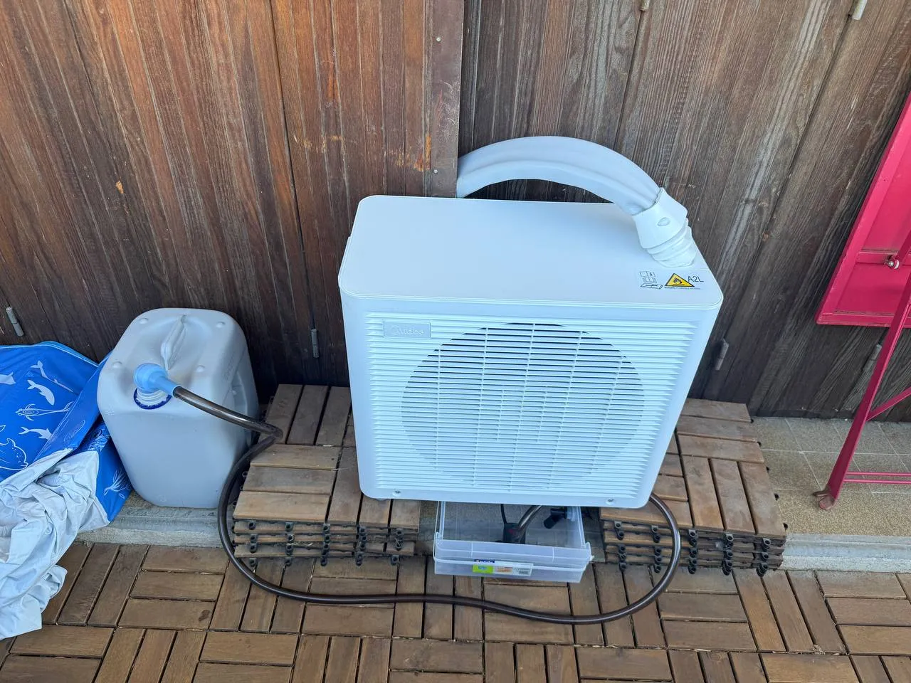 Climatiseur monobloc Midea posé sur une terrasse en bois. Un bac transparent est glissé sous l’appareil et un tuyau souple rejoint un jerrican gris posé à gauche.