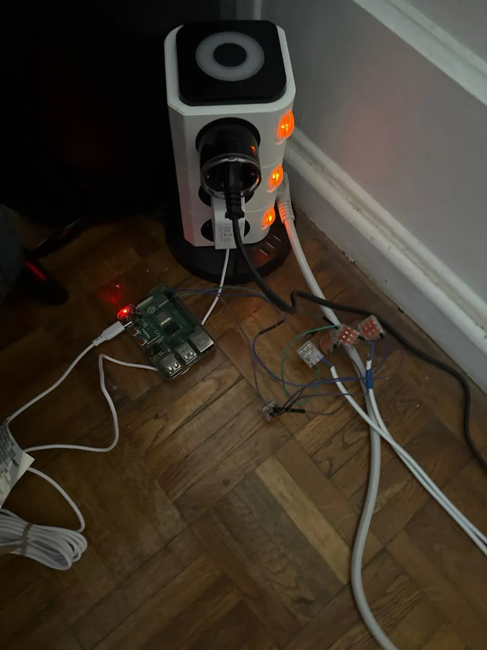 Raspberry Pi posé au sol, relié par des câbles à des bornes de connexion rapide, devant une multiprise verticale dont les prises sont allumées.